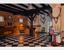 Wohnraum im rustikalen Stil mit schwarz-weiß kariertem Fliesenboden, freiliegenden Ziegel- und Steinwänden sowie dunklen Holzbalken. Ein Kamin mit Feuer, Jagdtrophäen an den Wänden, antike Möbel und eine rote Schneidemaschine prägen das Interieur.