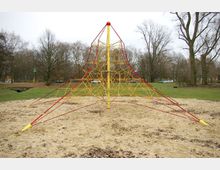 Klettergerüst in Form einer Pyramide mit gelben und roten Seilen auf einem Sanduntergrund, umgeben von einer Wiese und kahlen Bäumen in einer Parkanlage. Im Hintergrund sind eine Straße und parkende Autos zu sehen.