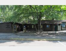 Holzgebäude mit Satteldach, umgeben von hohen Bäumen, im Vordergrund ein Gehweg, eine kleine Verkehrsampel und ein Parkplatzschild; links ist eine Fahrradstation erkennbar.