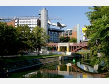Moderne Stadtansicht mit dem Deutschen Technikmuseum in Berlin, gekennzeichnet durch ein auf einer Plattform ausgestelltes Flugzeug. Im Vordergrund ein Kanal mit Uferbefestigungen und grüner Vegetation, eine rote Brücke und ein Gebäudekomplex im Hintergrund.