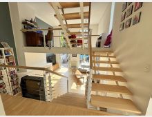 Blick in einen modernen, offenen Wohnraum mit Galerieebene und freitragender Holztreppe mit Metallgeländer. Unten sind ein Wohnzimmer mit Sitzmöbeln, Bücherregal und großen Glas-/Terrassentüren zu sehen, oben eine offene Arbeits- oder Musikfläche mit Möbeln und Instrumenten.