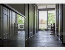 Ein moderner Flur mit dunklem Holzboden, beidseitig schwarzen Schiebetüren und einer Fensterfront am Ende, die auf einen Balkon mit Bäumen im Hintergrund blickt; links und rechts stehen dekorative Holzfiguren, daneben ein weißer Designstuhl und eine kleine Pflanze.