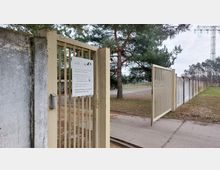 Ein geöffneter, gelber Metallzaun in einer umzäunten Außenanlage, flankiert von einer verwitterten Betonmauer. Im Hintergrund sind Bäume, ein Strommast und ein asphaltierter Weg sichtbar.