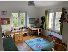 Helles Kinderzimmer mit zwei Fenstern, einem Schreibtisch, einer Weltkarte, einem Globus und Bastelmaterialien. Der Raum hat einen Teppich mit Meeresmotiven, Holzboden und Korbaufbewahrung.
