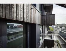 Moderne Wohngebäude mit einer Fassade aus dunklem Holz und großen Fenstern; Balkone mit schwarzen Metallgeländern ragen hervor. Im Hintergrund sind weitere moderne Gebäude und ein gepflasterter Innenhof sichtbar.
