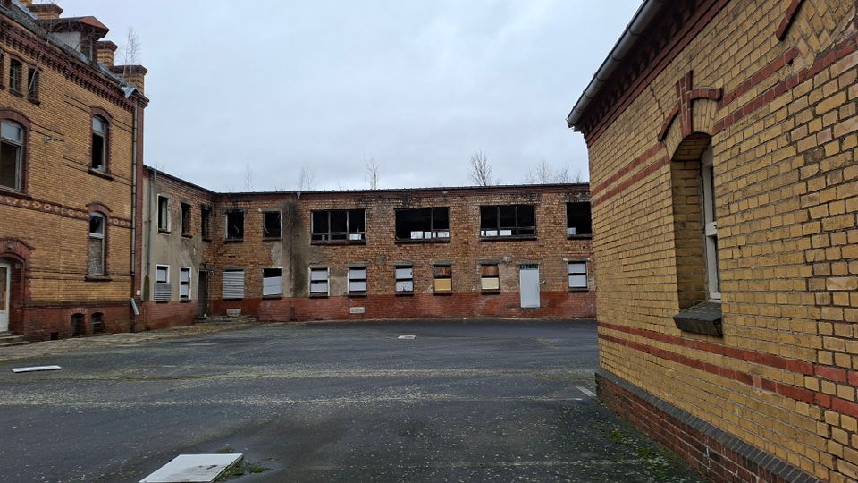 Innenhof eines verlassenen Backstein-Industriekomplexes mit mehrstöckigen Gebäudefassaden, teils vernagelten oder zerstörten Fenstern und dunklen Öffnungen. Der asphaltierte Hof ist weitgehend leer, umgeben von gelb-rotem Mauerwerk unter grauem Himmel.