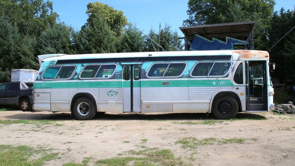 Ein alter, rostiger Linienbus in grün-weißer Lackierung steht auf einem gras- und erdbedeckten Platz, umgeben von Bäumen im Hintergrund. Rechts neben dem Bus ist die geöffnete Vordertür sichtbar, während links ein geparktes Auto und ein Anhänger stehen.
