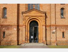 Fassade einer Kirche aus gelbbraunem Ziegelstein mit einem großen Rundbogenportal. Über dem Eingang verläuft ein verziertes Gesims mit Skulpturen auf beiden Seiten, und durch die geöffnete Tür sind Personen sichtbar.