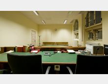 Ein einfacher Gerichtssaal mit einem grünen Richtertisch im Vordergrund, auf dem Bücher, Akten und Computergeräte stehen. Im Hintergrund befinden sich Holzbänke, ein Mantelständer, eine Standuhr sowie hohe Fenster mit Gitterelementen.