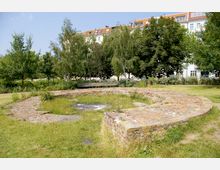 Eine Grünfläche mit Mauerresten aus Ziegelsteinen, die einen runden Grundriss andeuten, umgeben von Bäumen und Rasen. Im Hintergrund sind Wohnhäuser mit roten Dächern und weißen Fassaden sichtbar.