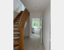 Helle Inneneinrichtung mit einem Flur, einer Holztreppe auf der linken Seite und einem offenen Zugang zu einem Wohnzimmer mit bodentiefen Fenstern und Blick auf einen Garten. Der Boden besteht aus hellem Laminat, und die Wände sind weiß gestrichen.