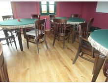 Ein Raum mit Holzfußboden, dunkelbraunen Stühlen und runden Tischen, die mit grünen Tischtüchern mit Fransen dekoriert sind; die Wände sind rot, und an einer Seite ist ein Fenster mit grünem Rahmen zu sehen.