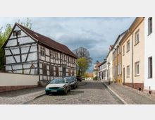 Straßenszene in einer kleinen historischen Stadt mit Kopfsteinpflaster, einem Fachwerkhaus links und modernen Wohnhäusern rechts. Im Hintergrund sind Bäume und ein Kirchturm zu sehen.