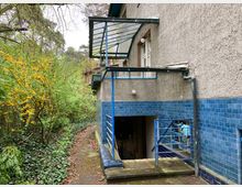 Seitenansicht eines Gebäudes mit verputzter, grauer Fassade und blau gefliester Sockelzone. Ein Metallgeländer und ein gläsernes Vordach befinden sich über einer Treppe, die zu einem Kellereingang führt. Umgeben von dichter Vegetation mit gelb blühenden Sträuchern und Efeu.