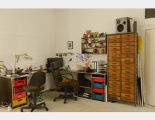 Ein Arbeitsraum mit zwei Schreibtischen, Bürostühlen und einem Computer. An der Wand befinden sich ein Regal mit Büchern und Gegenständen sowie ein hoher Schrank mit zahlreichen kleinen Holzschubladen; der Boden ist aus hellem Material.