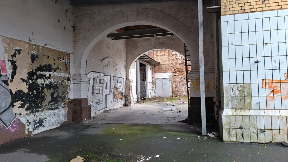 Verlassene Industriehalle mit großen gemauerten Rundbögen, abblätterndem Putz und Graffiti an den Wänden. Rechts sind weiß gekachelte Wandflächen zu sehen; im Hintergrund führen die Bögen in einen offenen Bereich mit freiliegendem Mauerwerk.
