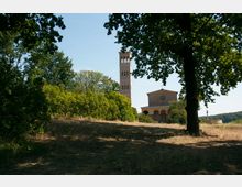 Historische Kirche mit einer Fassade aus Ziegelstein und einem hohen Glockenturm, umgeben von Bäumen und Büschen auf einer leicht bewachsenen Wiese unter klarem Himmel. Im Vordergrund werfen große Bäume Schatten auf den Boden.