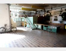 Industriell gestalteter Innenraum mit freiliegenden Rohren, einer Metalltreppe, vintage Möbeln und Pflanzen. Ein Fahrrad steht neben einer weißen Wand, und Holzböden dominieren den Raum.