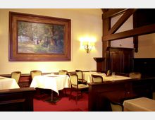 Restaurant-Innenraum mit klassischem Holzinterieur, roten Teppichboden und gedeckten Tischen mit weißen Tischdecken. An der Wand hängt ein großes Landschaftsgemälde neben einer stilvollen Wandleuchte.