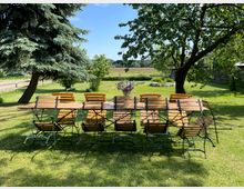 Langer Holzgartentisch mit mehreren Klappstühlen auf einer gepflegten Rasenfläche unter großen Bäumen. Im Hintergrund sind ein ländlicher Garten mit Sträuchern und Wiesen sowie Felder am Horizont zu sehen.
