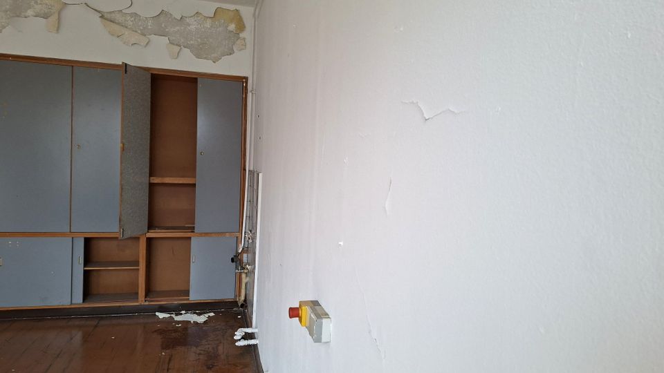 Leerer Innenraum mit abblätternder Deckenfarbe und stark abgenutztem Holzboden; an der linken Wand steht ein eingebauter Schrank mit teils offenen Türen und Regalböden. An der rechten Wand befinden sich Risse im Putz sowie ein Not-Aus-Schalter.