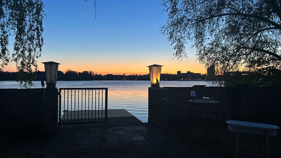 Blick von einer kleinen Uferterrasse mit Geländer und zwei Laternen auf einen ruhigen See bei Sonnenuntergang. Im Vordergrund rahmen Bäume und eine niedrige Mauer den Zugang zu einem kurzen Steg; am Horizont sind Silhouetten von Gebäuden zu sehen.