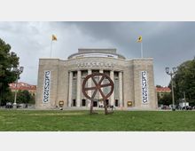 Die Volksbühne in Berlin, ein Theatergebäude mit neoklassizistischer Architektur, geprägt durch eine Fassade mit Säulen und großen rechteckigen Fenstern. Im Vordergrund steht eine moderne Metallskulptur auf einer Grünfläche, umgeben von Bäumen und Straßenlaternen.
