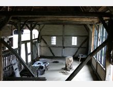 Innenraum eines Fachwerkgebäudes mit Holzbalken, weiß verputzten Wänden und zwei kleinen Fenstern mit Sprossenglas. Der Raum ist spärlich möbliert, mit einem Holzbock, Werkzeug und anderen Gegenständen auf dem Boden.