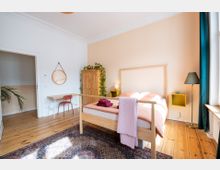 Helles Schlafzimmer mit einem Holzbett, einem rosa Bezug und daran hängenden Handtüchern. Der Raum enthält einen Holzkleiderschrank, einen Schreibtisch mit rotem Stuhl, eine Stehlampe, ein Fenster mit grünen Vorhängen und einen Teppich auf Holzboden.