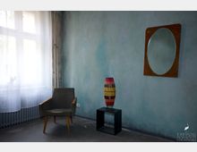 Ein minimalistisches Zimmer mit einer blassblauen Wand, einem großen Fenster mit transparentem Vorhang, einem grauen Sessel, einem kleinen schwarzen Beistelltisch mit einer farbigen Lampe und einem ovalen Wandspiegel.