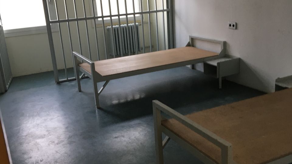 Spartanischer Innenraum mit zwei einfachen Metallbetten und kleinem Nachttisch auf dunklem Boden. Am Fenster befindet sich ein Gitter mit Tür, dahinter Heizkörper und Tageslicht; an der Wand ist eine Leuchtstofflampe montiert.