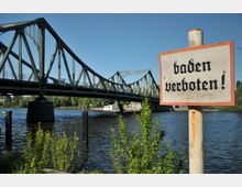 Eine grüne Stahlbrücke überspannt einen Fluss, umgeben von Bäumen und Vegetation; im Vordergrund steht ein Schild mit der Aufschrift "Baden verboten!".