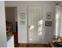 Flur in einem Wohnhaus mit einer weißen Tür in der Mitte, flankiert von vier gerahmten Bildern an der Wand. Links führt ein offener Durchgang in ein anderes Zimmer mit Regalen, und rechts befindet sich ein Fenster mit weißen Vorhängen, durch das Tageslicht einfällt. Im Vordergrund ist eine Ablage mit Spielsachen erkennbar.