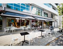 Straßenansicht eines modernen Gebäudes mit großen Fenstern, vor dem ein Café mit weißen Tischen und Stühlen auf einer gepflasterten Fläche eingerichtet ist. Eine Markise mit der Aufschrift "world of gaming" überspannt den Bereich, und ein beleuchteter Innenraum ist durch die Fenster sichtbar.