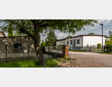 Wohngebiet mit einem flachen, weiß gestrichenen Gebäude und Vorgarten, umgeben von einem metallenen Zaun. Im Vordergrund ein gepflasterter Weg, ein Baum mit grüner Krone und ein Zaunstück mit Backsteinsäulen.