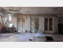 Ein verlassener, verfallener Raum mit beschädigten Wänden, zerbrochenen Türen mit Lamellen aus Holz und einer kaputten Decke, durch die Licht einfällt. Der Boden ist mit Schutt und Unrat bedeckt, ein alter Heizkörper steht an der linken Wand.