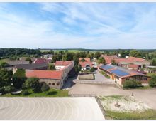 Luftaufnahme eines ländlichen Hofgeländes mit mehreren Gebäuden mit roten Dächern, darunter moderne und traditionelle Bauwerke. Im Vordergrund ein großer Reitplatz mit hellem Sand, im Hintergrund Wiesen und Wälder.