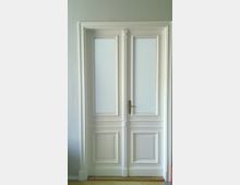 Weiße Doppeltür im klassischen Stil mit dekorativen Kassetten und schlichter Verglasung im oberen Teil, umgeben von einer beige gestrichenen Wand.