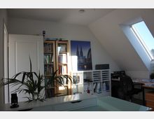 Ein Büro in einem Dachgeschoss mit Schreibtisch, Bücherregalen und einem Drucker. Auf der Wand hängt ein Bild des Kölner Doms, daneben ein großes Dachfenster, das viel Tageslicht hereinlässt. Im Vordergrund steht eine Zimmerpflanze auf einer Kommode.