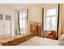 Helles Schlafzimmer mit großen Doppelfenstern, die von cremefarbenen Vorhängen umrahmt sind. Der Raum enthält ein Holzbett, einen roten Sessel, einen kleinen Glastisch mit Stuhl und eine dekorative Trennwand aus Holz.