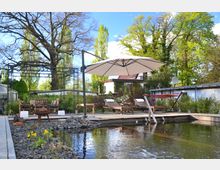 Gartenanlage mit einem Teich im Vordergrund, umgeben von Kies und Pflanzen. Im Hintergrund befinden sich Holzliegen, ein großer Sonnenschirm, eine Hängematte sowie ein Essbereich mit Holzmöbeln; dahinter sind Bäume und ein Haus sichtbar.