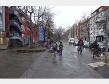 Städtische Straßenszene mit einer Gehwegfläche, mehreren Fahrrädern an einem Fahrradständer und Personen, die gehen oder Fahrrad fahren; im Hintergrund bunte Wohngebäude, graffitibedeckte Wände und parkende Autos entlang der Straße. Ein Straßenschild mit der Aufschrift „Gitschiner Straße“ ist sichtbar.