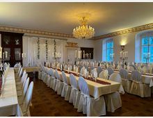 Eleganter Festsaal mit langen, festlich gedeckten Tischen, weiß eingedeckten Stühlen und Kerzenleuchtern. Ein prunkvoller Kronleuchter hängt von der Decke, umgeben von Stuckverzierungen und großen Fenstern mit Blick nach draußen.