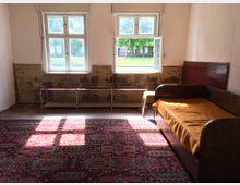 Ein schlicht eingerichtetes Zimmer mit abgenutzten Wänden, zwei Fenstern, von denen eines geöffnet ist, einer Holzbank unter den Fenstern und einer Holzliege mit orangefarbener Decke rechts. Der Boden ist mit einem großen, gemusterten Teppich bedeckt, und Tageslicht fällt durch die Fenster ins Zimmer.