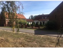 Straßenszene in einem Dorf mit mehreren Häusern im traditionellen Stil, roten Ziegeldächern und Zäunen aus Holz. Im Vordergrund führt ein asphaltierter Weg an einer Wiese vorbei, und ein Baum mit herabhängenden Zweigen rahmt das Bild ein.