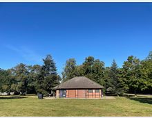 Ein einstöckiges Gebäude mit einem dunklen Ziegeldach steht inmitten eines grünen Parks, umgeben von hohen Bäumen und einer Rasenfläche unter einem klaren blauen Himmel.