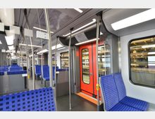 Innenraum einer modernen U-Bahn mit blauen Sitzen und regelmäßigen Mustern. Sichtbar sind Haltestangen aus Metall, eine rote Tür mit Sichtfenster und ein daneben liegendes Fenster mit Blick auf ein Lager.