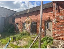 Hinterhof mit einer alten, roten Ziegelsteinfassade, mehreren kleinen Fenstern und einer Holztür. Der Hof ist überwuchert mit Gras und Pflanzen, während ein Holzgestell und Dekorationen im Vordergrund sichtbar sind.