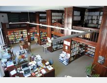 Innenraum einer Bibliothek mit zwei Ebenen, geprägt von dunklen Holzregalen, die Bücher und Ordner enthalten. In der Mitte stehen Schreibtische mit Büromaterial sowie ein kleiner Sitzbereich mit Tisch und Stühlen, umgeben von zahlreichen Regalen.