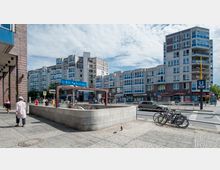 Eingangsbereich der U-Bahn-Station "Pankstraße" in einer städtischen Umgebung, umgeben von mehrstöckigen Wohngebäuden mit modernen Fassaden und Geschäften im Erdgeschoss. Mehrere Fahrräder stehen am Rand, während Fußgänger und Fahrzeuge die Szene beleben.